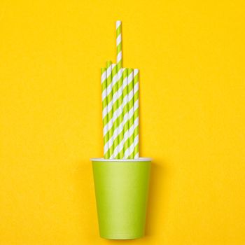 Multicolored cocktail tubes in green paper disposable cup on a yellow background