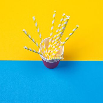 a bundle of multi-colored drinking straws in a paper Cup on a yellow and blue background. fashion minimal. flat lay