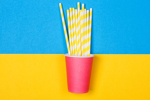 a bundle of multi-colored drinking straws in a paper Cup on a yellow and blue background. fashion minimal. flat lay