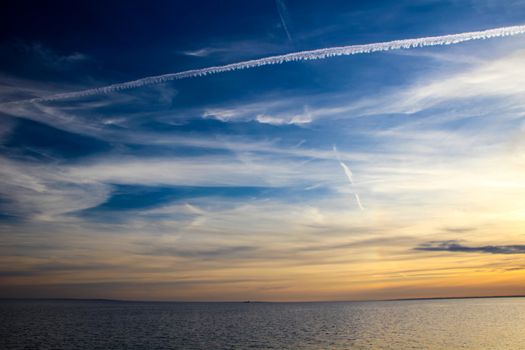 Chemtrails and clouds on a blue and orange sky in spring at the sea.