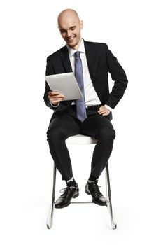 Cheerful young businessman sitting on a chair and looking at digital tablet. Isolated on white background.