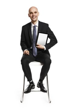 Cheerful young businessman sitting on a chair and showing digital tablet. Isolated on white background.