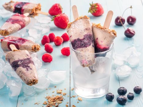 Homemade popsicle and cherry, blueberry, strawberry, raspberry on blue wooden background. Healthy summer breakfast. Popsicles from yogurt and banana with berries, granola and chia seeds