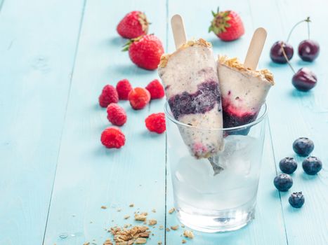 Homemade popsicle and cherry, blueberry, strawberry, raspberry on blue wooden background. Healthy summer breakfast. Popsicles from yogurt and banana with berries, granola and chia seeds. Copy space.