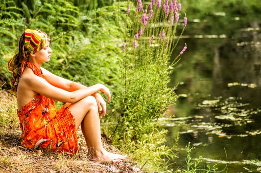 a woman with dreadlocks is sitting on the shore of the pond looking far away