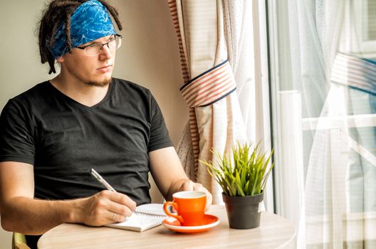 a young man with dreadlocks is writing in a notebook coffee at home