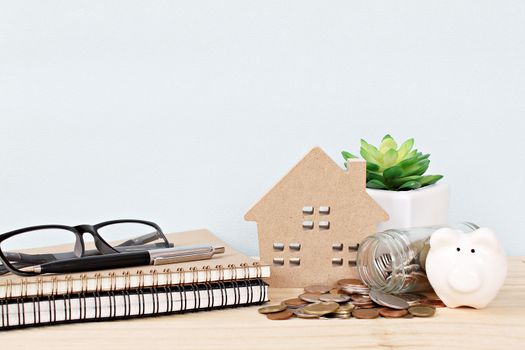 Business, finance, savings, property ladder, mortgage or loan concept : Wood house model, piggy bank, coins scattered from glass jar, notebooks, pen and eyeglasses on wooden background