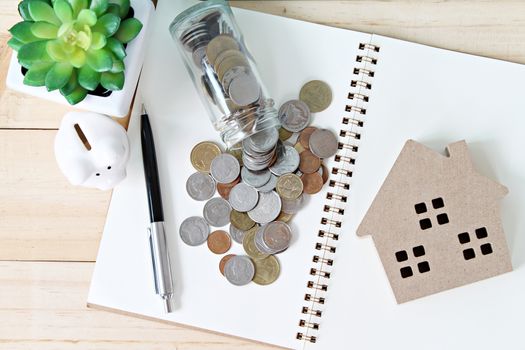 Business, finance, saving money, banking, property loan or mortgage concept : Top view or flat lay of wood house model, coins, open notebook paper and piggy bank on office desk table