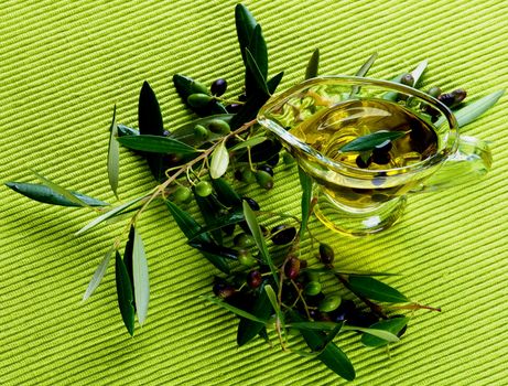 Fresh Olive Oil in Glass Gravy with Raw Green and Black Olives with Leafs on Green Textile background. Top View