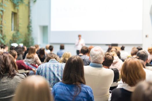 Business and entrepreneurship symposium. Female speaker giving a talk at business meeting. Audience in conference hall. Rear view of unrecognized participant in audience. Copy space on whitescreen.
