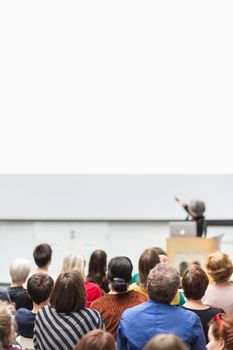 Business and entrepreneurship symposium. Female speaker giving a talk at business meeting. Audience in conference hall. Rear view of unrecognized participant in audience. Copy space on whitescreen.