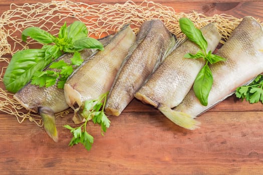 Several uncooked carcasses of the notothenia fish without of a heads and tails and twigs of basil and parsley closeup on the fishing net on a dark wooden surface
