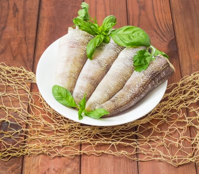 Carcasses of the notothenia fish without of a heads and tails and with peeled scales and prepared for cooking, basil and parsley twigs on the white dish on a dark wooden surface with fishing net
