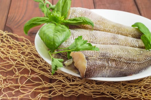 Fragment of a white dish with carcasses of the notothenia fish without of a heads and tails and with peeled scales, basil and parsley twigs on a dark wooden surface with fishing net closeup
