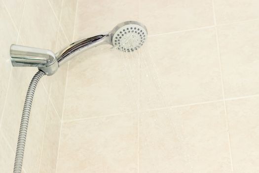 Water flowing from shower head with turn lever of a spray settings and metal shower hose in holder on a background of a wall with beige tiles
