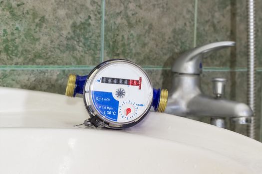 Not connected residential water meter for consumption measuring of a cold water on a wash basin on background of a handle mixer tap and wall with green tiles
