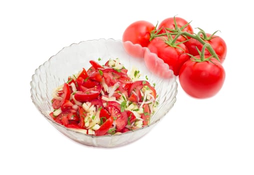 Vegetable salad of the fresh sliced tomatoes, white bell pepper, onion and potherb in a glass salad bowl against of the cluster of fresh tomatoes on a white background
