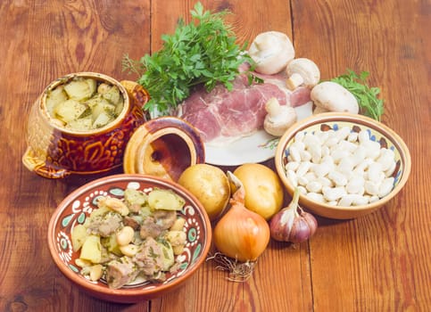 Ukrainian version of the dish Chanakhi in a clay bowl - potatoes with meat, mushrooms and haricot beans baked in a clay pot and ingredients for its preparation on a surface of an old wooden planks
