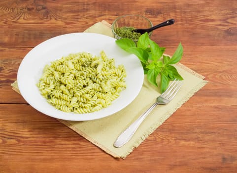 Cooked spiral pasta with sauce pesto on the white dish, pesto in small glass bowl with spoon, fork and basil twig on a napkin on a surface of old wooden planks
