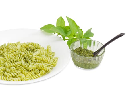 Fragment of the white dish with cooked spiral pasta with sauce pesto, pesto in small glass bowl with spoon beside and basil twig on a white background
