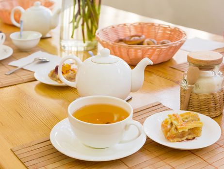 White cup with a herbal tea and a piece of cake on a saucer on the bamboo napkin on a background of a teapot and other tea appliances
