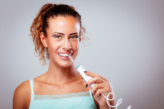 Smiling young woman with braces cleaning her teeth with water flosser. Looking at camera.