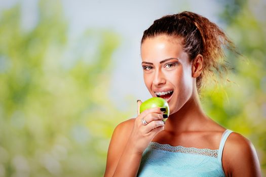 Smiling young woman with braces on white teeth holding green apple and looking at camera.