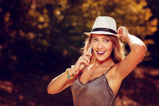 Beautiful smiling young woman with summer hat enjoying in the nature in autumn and talking on smartphone.
