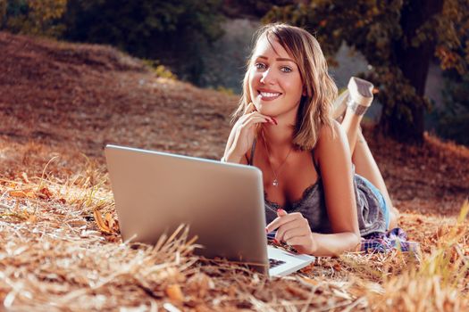 Cute smiling young woman lying on the withered grasses in autumn and using laptop.