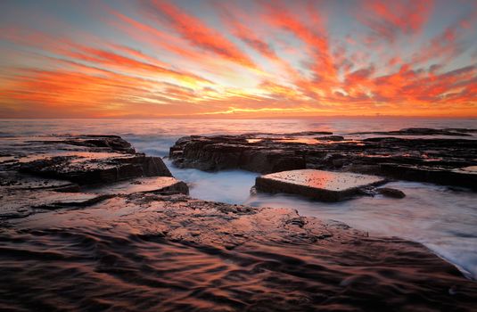 Red sunrise skies over the ocean and eroded rock channel and reclections on the dark wet rocks.  Location North Narrabeen, northern beaches Sydney Australia, a beautiful travel destination.