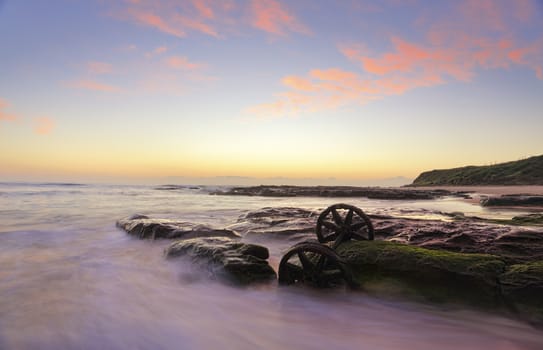 Lovely autumn sunrise over Windang Beach with one of the many abandoned rusty train wheels exposed on mossy rocks at low tide.