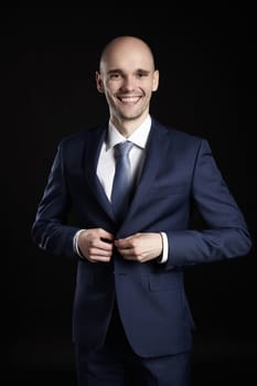 Portrait of young businessman fastens his jacket. Studio shot on black background.