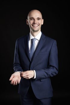 Portrait of smiling man fixing cuffs his suit. Studio shot of black background.