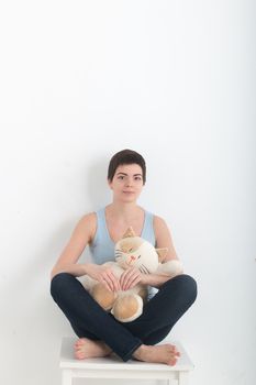 Young attractive smiling woman practicing yoga, sitting in Half Lotus exercise Ardha Padmasana pose, working out wearing dark bleu jeans indoor full length, white plush toy cat on her lap.