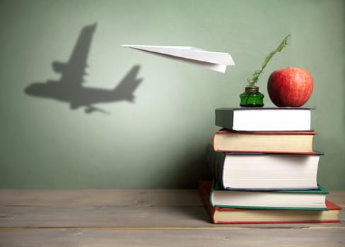 Paper plane with shadow of an aircraft next to a stack of books, quill and apple