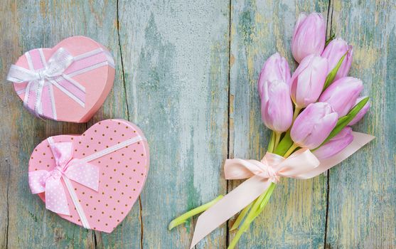 Bouquet of pink tulips tied with a satin ribbon and two gift boxes in the shape of a heart with a bow, on the old blue wooden background, with space for text