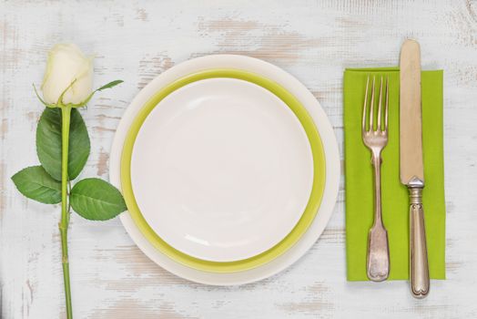 White and green empty plates, green linen napkin, old fork and knife and white rose flower on an old wooden table; top view, flat lay, overhead view