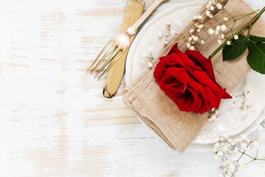 White plates, linen napkin, old fork and knife, and red rose flower on an old wooden table; top view, flat lay, overhead view