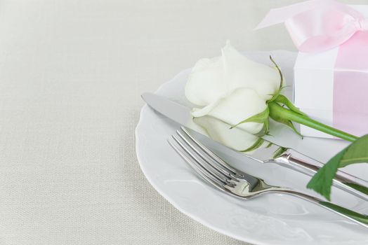 Beautiful decorated table with white plates, gift box with a pink ribbon, cutlery and white rose flower on tablecloths, with space for text