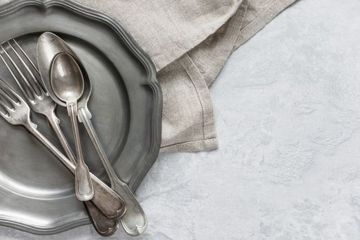 Various silverware on a pewter plate and gray flax napkin are on the background of gray concrete surface, with copy-space