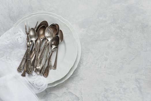 Various silverware on a porcelain plate and white napkin with Belgian lace are on the background of gray concrete surface, with copy-space