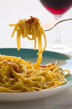 Closeup of spaghetti carbonara on fork with egg, smoked bacon and cheese over a table with a red wine glass