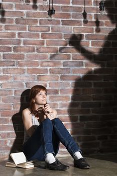 Caucasian girl sitting on the floor reading a horror or scary thriller book. Dramatic lighting with girls eyes wide open and long shadows of woman with knife on the wall near her. Afraid lady looking to somebody. Terrified expression