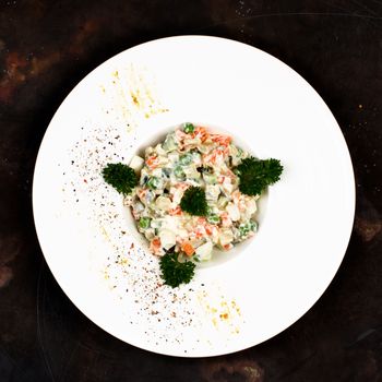 Traditional Russian Salad Olivier with Boiled Chicken Meat, Eggs and Vegetables Decorated with Parsley in White Plate on Dark background. Top View 