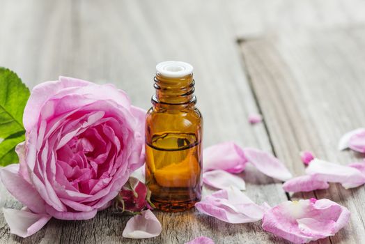 Dark glass vial with rose essential oil and flower of pink rose on a wooden background