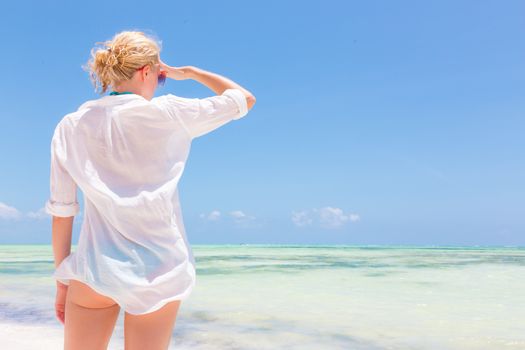 Happy woman enjoying, relaxing joyfully in summer by tropical blue water. Beautiful caucasian model wearing white beach tunic on vacations looking down picture perfect Paje beach, Zanzibar, Tanzania.