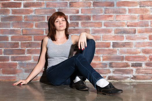 Attractive young woman in casual clothes, blue jeans and vest grey is looking at camera and smiling sitting on a floor by the brick wall. Hipster girl. Youth fashion, beauty. Red hair, haircare. Horizontal.