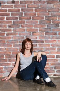 Attractive young woman in casual clothes, blue jeans and vest grey is looking at camera and smiling sitting on a floor by the brick wall. Hipster girl. Youth fashion, beauty. Red hair, haircare. Vertical.