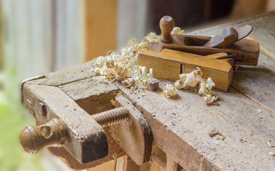 Fragment of old woodworking workbench with planing stop and shoulder vise with wooden screw, two wooden hand planes lying on workbench among a shavings
