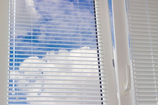 Fragment of the modern plastic window with Venetian blinds and view of the sky with clouds across slats of a window blind
